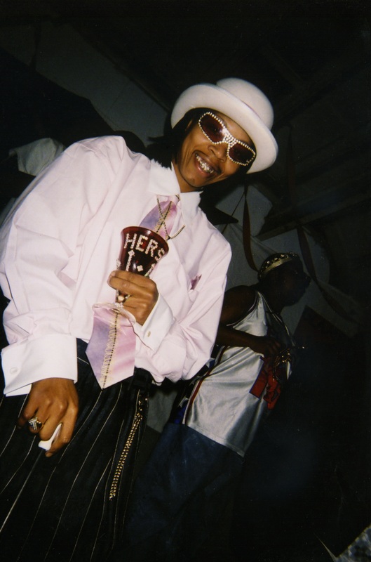 A masculine performer at Soakie's smiles while holding a chalice with gems spelling "HERS". 